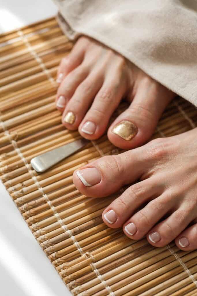French tip summer toe nails with gold glitter accent on a bamboo mat — minimalist and elegant pedicure with clean natural tones.
