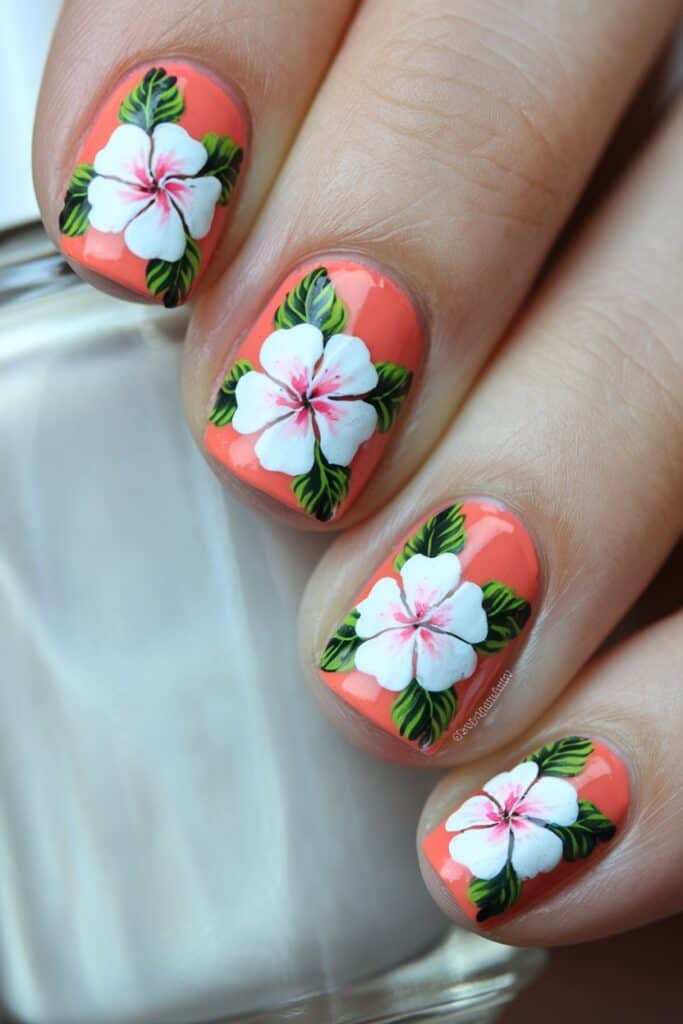 Short coral acrylic nails with white tropical hibiscus flower nail art and green leaves.