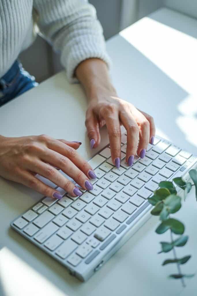 Person with lavender nail polish typing on a white keyboard with a small plant nearby.
