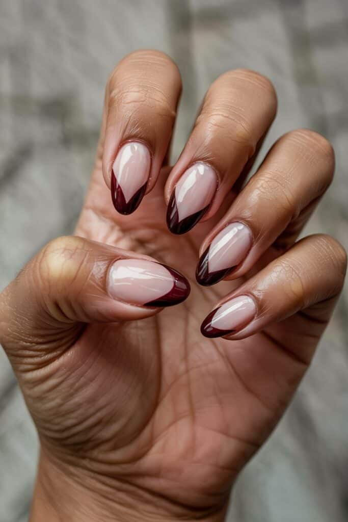Almond-shaped nails with a glossy nude base and deep burgundy diagonal French tips. Hands are held in a relaxed, slightly curled pose.