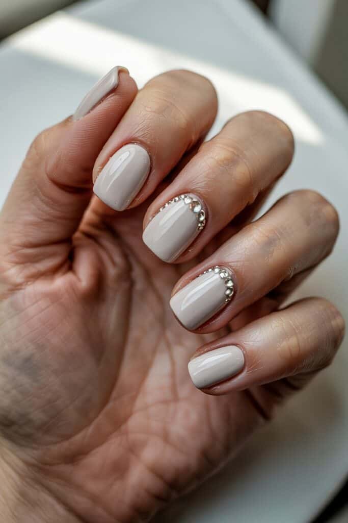 Short glossy nude nails featuring a curved row of silver rhinestones along the base of two accent nails. Close-up shows the shimmer and smooth finish.
