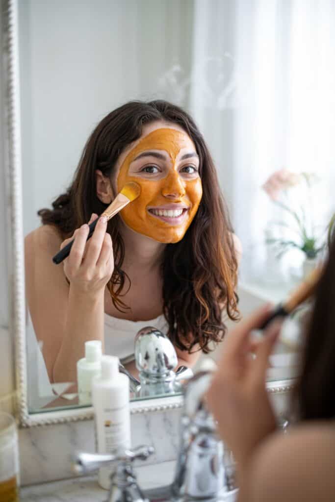 Woman applying golden turmeric face mask evenly across her face with fingertips.
