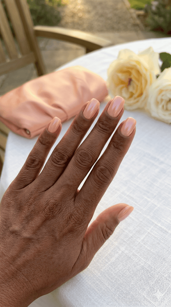 Close-up of mature hands with deep skin tone featuring glazed peach pearlescent nails, soft squoval shape, resting on a white linen table.