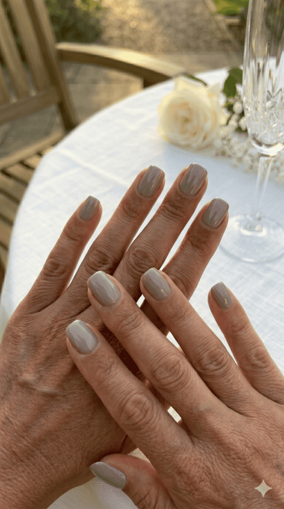Two hands with mature skin crossing fingers on a white linen table, featuring flawless high-gloss taupe lacquer nails, "Rich & Boogie" aesthetic.