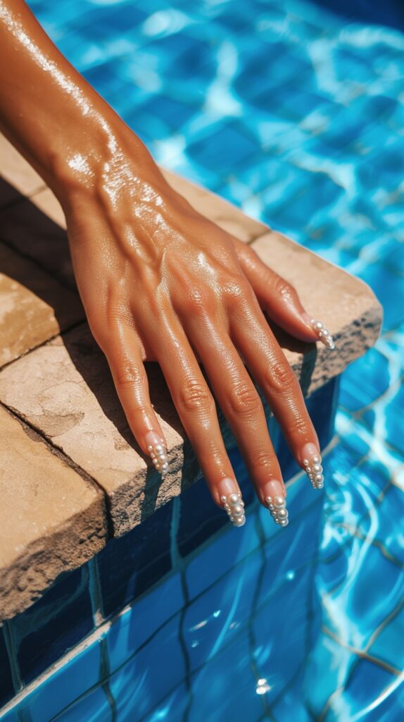 Tanned hand resting on a sunny pool ledge with long stiletto nails featuring tiny pearl bead accents at the tips, bright blue pool water reflecting in the background

