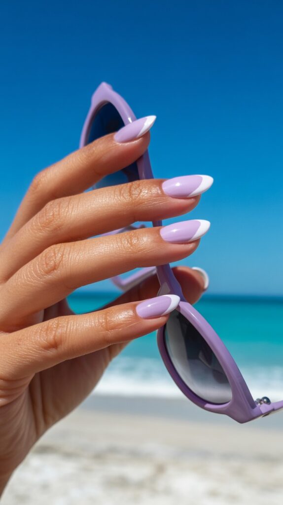 Hand holding lavender sunglasses at the beach with almond-shaped nails featuring a lavender and white micro French tip, bright blue ocean in the background

