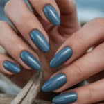 Beautiful blue manicure with oval-shaped nails against a beach background.